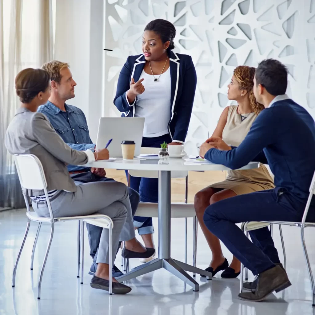 5 Personas en una reunión y una de ellas está de pie hablándole a los demás.